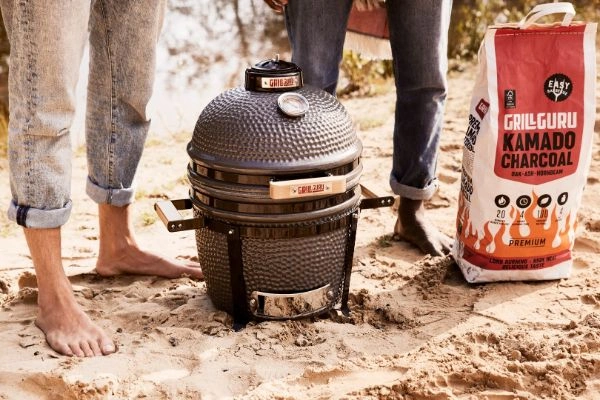 Gratar Grill Guru Original Compact pe nisip cu sac de carbuni langa el.